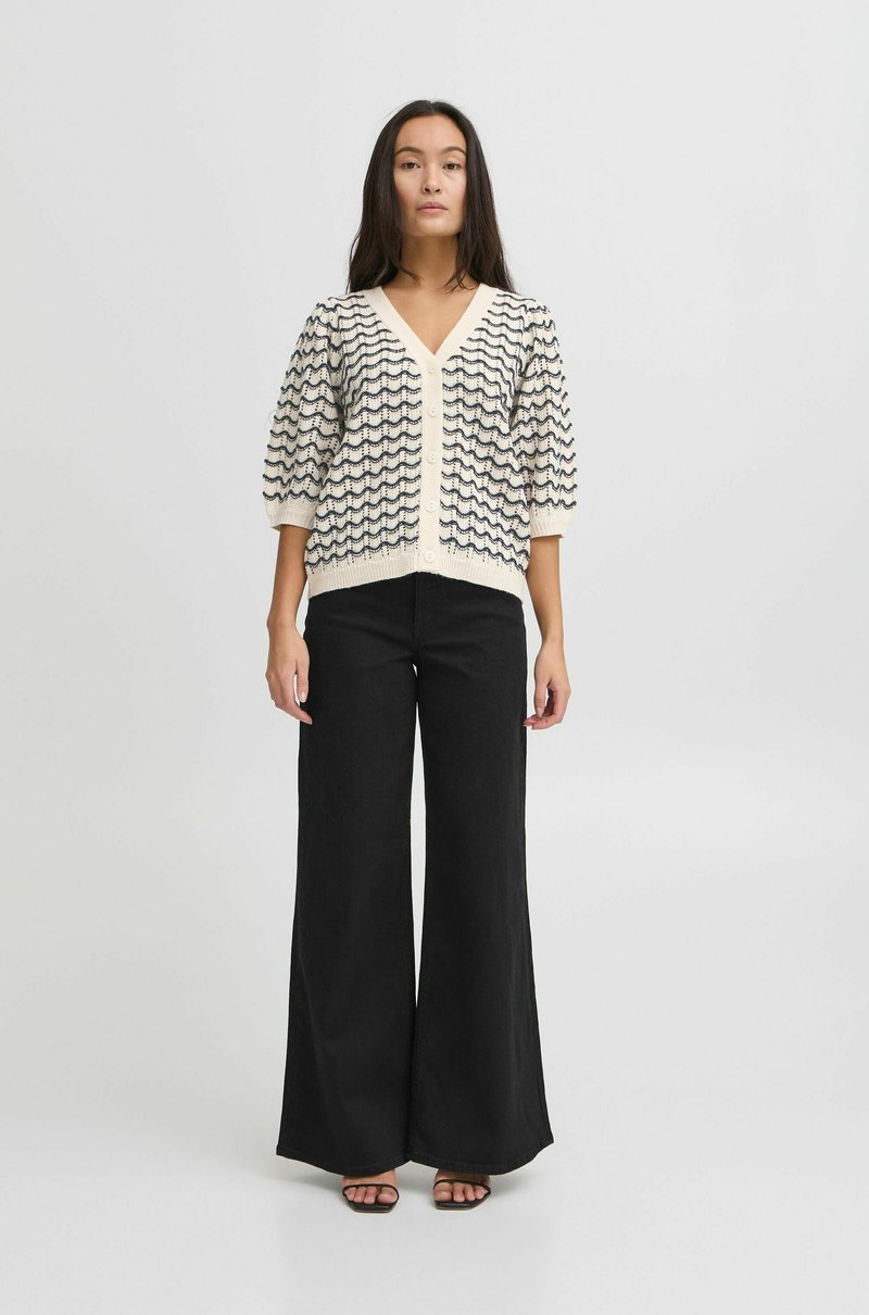 Cardigan en tricot crème avec un motif de vagues bleu marine, décolleté en V et fermeture à boutons. Assorti à un pantalon large taille haute noir et des sandales.