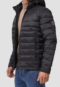 Veste matelassée noire avec capuche, dotée d'une fermeture éclair frontale, de poches latérales zippées et d'un patch logo sur la manche gauche. Tissu texturé.