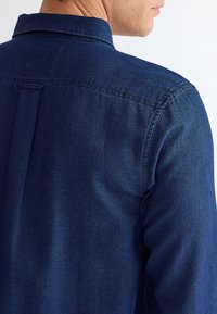 Uomo che indossa una camicia di denim blu scuro, visibile da dietro e parzialmente di lato, con dettagli visibili delle cuciture di collo e spalla.