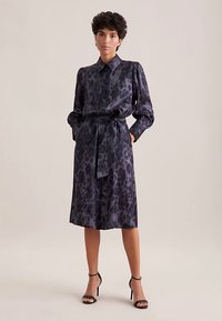 Donkere blousejurk met patroon, een kraag, lange mouwen en een ceintuur in de taille. Materiaal heeft een glanzende afwerking; voorzien van een luipaardprint.