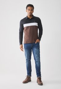 Man wearing a color-blocked long-sleeve shirt with black, white, and brown sections, blue jeans, and brown lace-up boots, standing with one hand in pocket.