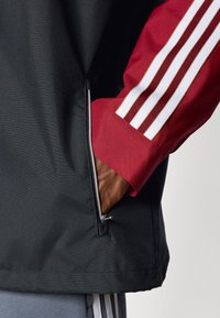 Svart jakke med røde ermer og tre hvite striper. Glidelåslomme på siden synlig, med en hånd inni. Myk stofftekstur.