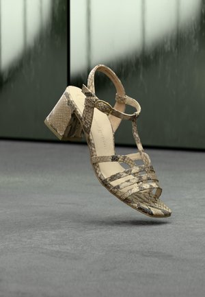 Sandale à talons hauts en peau de serpent beige avec bride à la cheville et boucle dorée, suspendue au-dessus d'un sol gris sur un fond sombre flou.