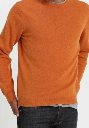 Homme portant un pull ras du cou orange par-dessus une chemise grise et un jean foncé, main reposant dans la poche, sur fond blanc.