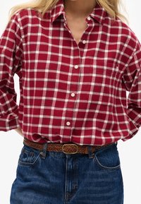 Camicia a quadri rossa e bianca in flanella con collo abbottonato, caratterizzata da una tasca sul petto e una cintura intrecciata marrone con fibbia dorata.