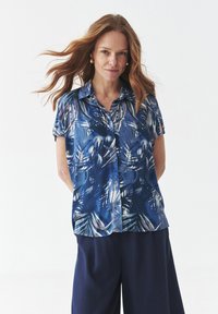 Camicia a maniche corte con bottoni, caratterizzata da un motivo di foglie tropicali blu su un tessuto leggero. Abbinata a pantaloni scuri e larghi.