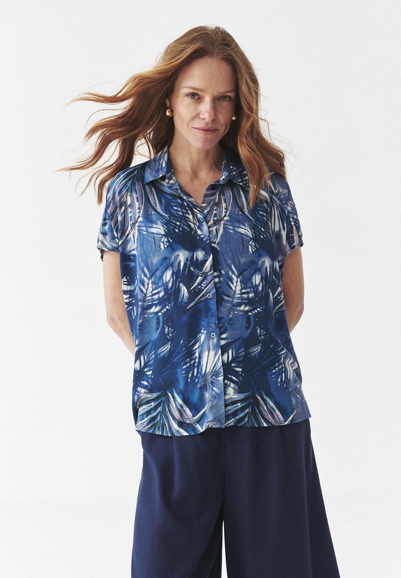 Camicia a maniche corte con bottoni, caratterizzata da un motivo di foglie tropicali blu su un tessuto leggero. Abbinata a pantaloni scuri e larghi.