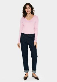 Lichtroze lange mouwen v-hals trui gecombineerd met donkere blauwe spijkerbroek, opgerolde mouwen en bruine platte schoenen. De outfit heeft eenvoudige stikdetails.