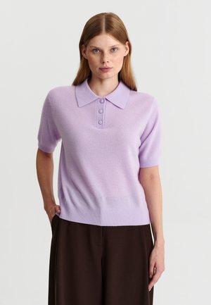 Femme aux cheveux roux raides portant un polo lavande à manches courtes et un pantalon marron foncé, debout devant un fond uni.