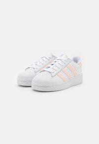 Vita Adidas Superstar-skor med ljusrosa rand och snäppa design, sedd i en vinkel på en vit bakgrund.