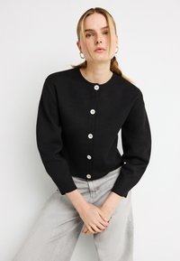 Μαύρη ζακέτα με στρογγυλή λαιμόκοψη, μακριά μανίκια και κλείσιμο με κουμπιά στο μπροστινό μέρος. Διακοσμείται με μεγάλα λευκά κουμπιά και υφασμάτινη πλεκτή υφή.