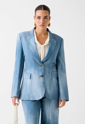 Vrouw met naar achteren gekamd haar draagt een vervaagde blauwe dubbelgeknoopte blazer, bijpassende broek, witte blouse, gouden oorringen en houdt een witte tas vast.