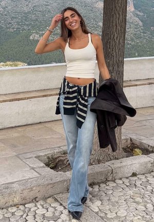 Weißes kurzes Tanktop, gestreiftes schwarz-weißes gebundenes Hemd, hellblaue ausgestellte Jeans, schwarze Jacke in der Hand, im Freien mit Bäumen im Hintergrund.