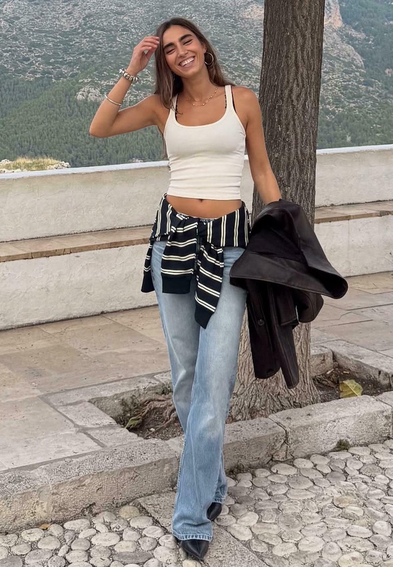 Witte cropped tanktop, gestreept zwart-wit knoopshirt, lichtblauwe wijde jeans, zwarte jas in de hand, buitenomgeving met bomen.