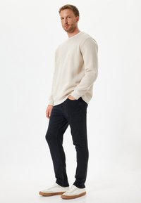 Sudadera beige, con puños acanalados, corte relajado; jeans oscuros, corte ajustado; zapatillas blancas con suelas marrón claro; textura de tela lisa.