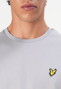 Pilkas crewneck marškinėliai su mažu geltonu ir juodu siuvinėtu erelio logotipu kairiajame krūtinės šone, pagaminti iš lygaus, tampraus audinio.