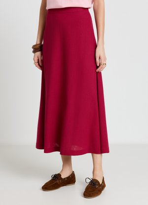 Femme portant une longue jupe rouge, des mocassins marron, un haut rose, des bracelets en bois et des bagues en or, debout devant un fond clair uni.