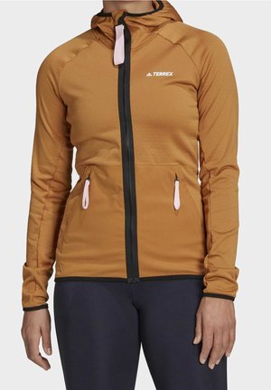 Kobieta w brązowej kurtce z kapturem na zamek błyskawiczny z różowymi suwakami i czarnych legginsach na jednolitym tle.