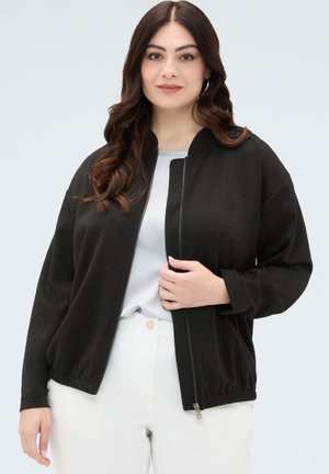 Vrouw met lang donker haar die een zwart jasje met ritssluiting draagt over een wit topje en een witte broek, staand tegen een egaal achtergrond.