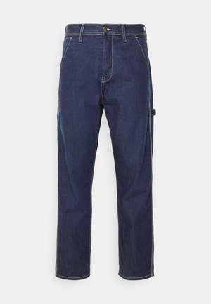 Mann trägt dunkelgraues kurzärmeliges T-Shirt mit Brusttasche, blaue locker sitzende Jeans und weiße Sneakers mit schwarzen Streifen, steht mit einer Hand in der Tasche.