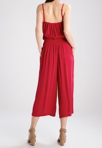 Mono rojo sin mangas con un cuerpo fruncido, diseño de pierna ancha y cintura elástica. Tejido suave, complementado por sandalias de tobillo beige.
