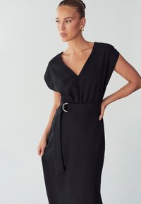 Musta mekko V-kaula-aukolla, lyhyillä hihoilla ja vyötäröstä vyötetty hopeisella soljella. Silkkinen kangas elegantilla ja minimalistisella muotoilulla.