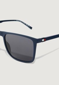 Óculos de sol retangulares mate na cor azul-marinho com lentes escuras e detalhes em vermelho, branco e preto na haste, em contraste com um fundo claro.