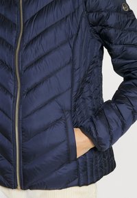 Mörkblå vadderad jacka med dragkedja fram, med chevron-sömmar och sidofickor. Mjukt, glänsande tyg med elastiska ärmslut och nederkant.