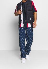 Džínová vesta s mnoha kapsami, námořnické a červené pruhované tričko, vzorované modré cargo kalhoty a bílé tenisky. Neformální, vrstvený outfit.