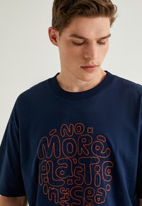 Marineblå bomulds t-shirt med orange broderet tekst, der siger "INGEN FLERE PLASTIKKER I HAVET", rund halsudskæring og korte ærmer.