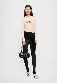 Fiorucci majica s dugim rukavima u svijetlo bež boji s crvenim logom, uparena s sjajnim crnim tajicama i cipelama s detaljem u obliku mašne srebrne boje, drži crnu torbicu.