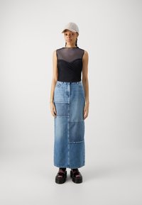 Svart ærmeløs topp med et gjennomsiktig lag, kombinert med et langt dongeri skjørt med lappverkdesign. Svart plattformstøvler og en lett caps.