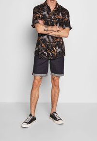 Camisa negra de manga corta con estampado tropical colorido, combinada con pantalones cortos de mezclilla oscura con dobladillos enrollados y zapatillas negras.