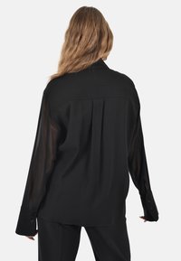 Maglia nera con retro in tessuto morbido e maniche in tessuto trasparente. Presenta un colletto, polsini con bottoni e un design plissettato sul retro.