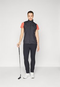 Black sleeveless vest over a red short-sleeve shirt, paired with navy fitted pants and white golf shoes. Golf club in hand.