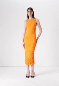 Robe sans manches orange avec fronces sur le côté, longueur genoux, en tissu extensible, assortie à des chaussures à talons hauts noires.
