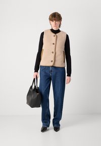 Μπεζ γιλέκο απ�ό φλις με καφέ τσέπες και κουμπιά, συνδυασμένο με σκούρα τζιν και μαύρες μυτερές γόβες, κρατώντας μια μαύρη τσάντα.