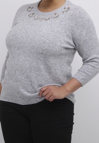 Pull gris en tissu doux, avec un col rond orné d'embellissements floraux décoratifs. Il a des manches longues et une coupe décontractée.