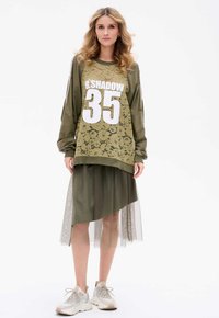 Olijfgroen oversized sweatshirt met kantoverlay en witte "35" afdruk, gecombineerd met een asymmetrische rok en lichte sneakers.