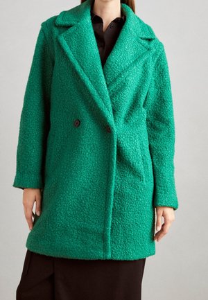 Kvinde iført en tekstureret grøn dobbeltradet frakke med brede revers over en mørk skjorte og bukser mod en ensfarvet baggrund.