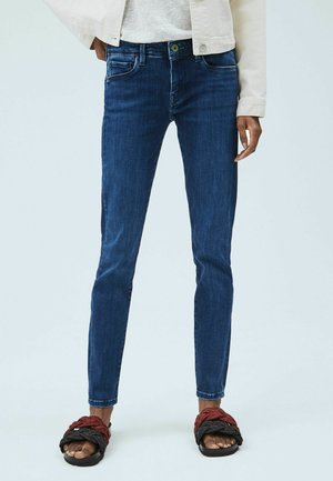 Person, die schlanke, dunkelblaue Jeans, eine cremefarbene Jacke und schwarze sowie rote geflochtene Sandalen trägt, steht vor einem hellgrauen Hintergrund.