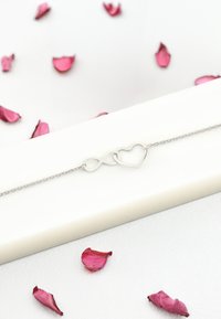 Silverarmband med en oändlighetssymbol kopplad till ett hjärtformad design. Lagt på en vit yta med spridda rosa rosblad.