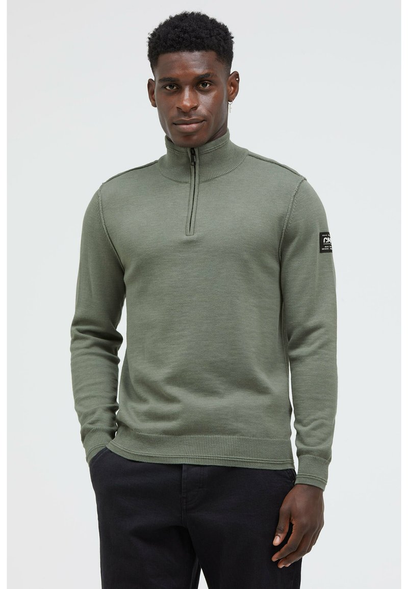 Olivengrüner Pullover aus Rippstrick mit einem Halbreißverschluss-Kragen und eng anliegenden Bündchen. Mit einem Patch auf dem linken Ärmel.