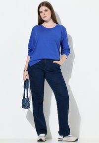 Niebieska bluzka z długim rękawem o luźnym kroju, zestawiona z ciemnymi dżinsami. Modelka trzyma małą niebieską torebkę. Look uzupełniają sneakersy z wielokolorowym wzorem.