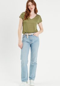 T-shirt verde oliva a V con maniche corte, abbinata a jeans azzurri dritti e una cintura con motivo, indossata con sneakers bianche.