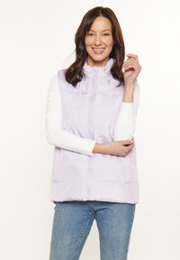 Vesta puf de culoare lavandă, cu fermoar frontal, guler înalt și design matlasat. Puritată deasupra unei bluze albe cu mânecă lungă și asortată cu blugi albaștri.