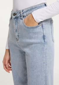 Jeans din denim de culoare albastră deschisă, cu talie înaltă, buzunare laterale și un detaliu subtil de decolorare. Materialul are o finisare netedă, ușor texturată.