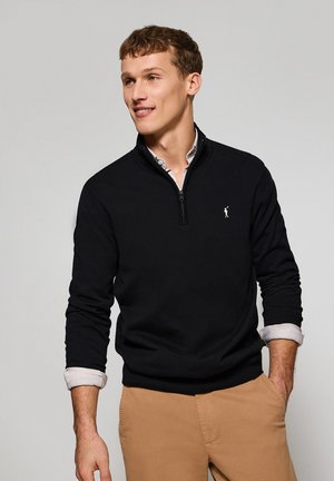 Schwarzer Quarter-Zip-Pullover mit gerippten Bündchen und Saum, ausgestattet mit einem kleinen weißen Logo auf der Brust. Getragen über einem hellen Hemd mit Kragen.
