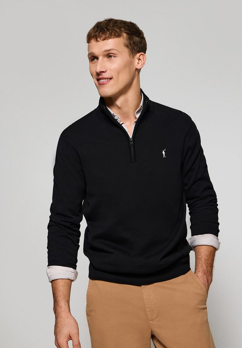 Svart kvartshigh zip-tröja med ribbad mudd och fåll, med en liten vit logotyp på bröstet. Bärs över en ljus skjorta med krage.