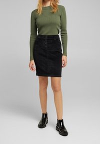 Femme portant un haut à manches longues vert olive, une jupe noire boutonnée au genou et des bottines noires brillantes, debout devant un fond gris.